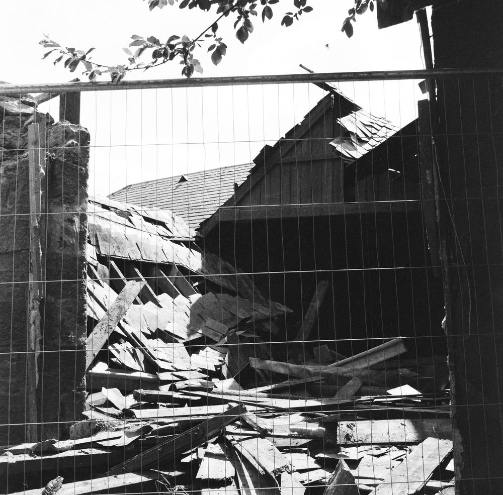 Photo looking through a mesh barrier at the remains of a collapsed farm building roof. The broken wooden rafters and slates are lying in a pile in the middle of the photo. In the background the remaining bits of roof are beginning to sag downwards.