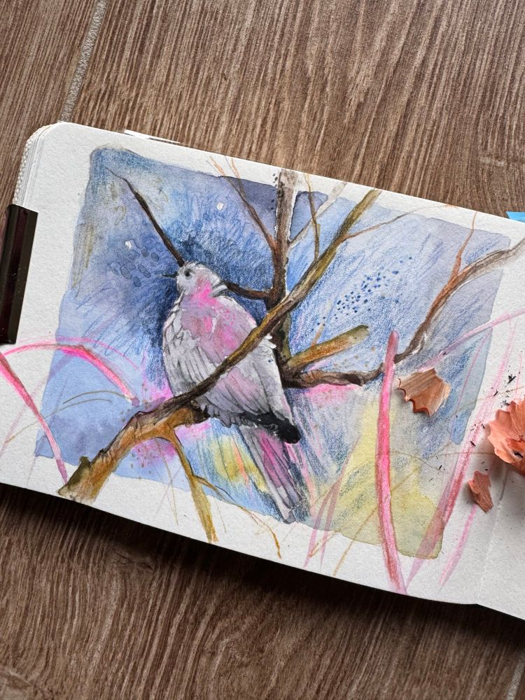 A watercolor illustration of a bird perched on a branch, surrounded by soft blue and pastel hues. The artwork features delicate details and a blend of colors, emphasizing the bird against a textured background. A wood surface is visible under the sketchbook.