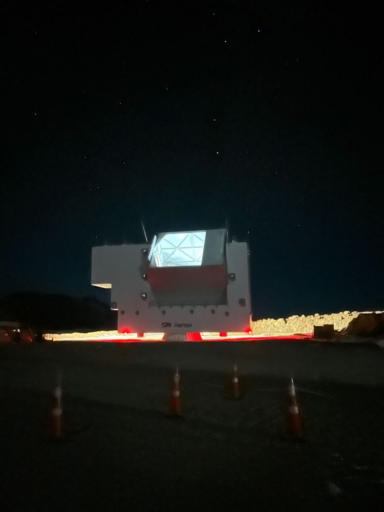 A night photo of a large building-sized telescope. The building is in two parts: a big u-shaped structure is mounted on a pivot, and itself ensnares a smaller brick-shaped box that can swivel with a hole in it. Some rocks in the background are visible in yellow, illuminated by an unseen source. Red safety lights at the bottom of the U element and ice blue light from within the telescope's opening complete the scene