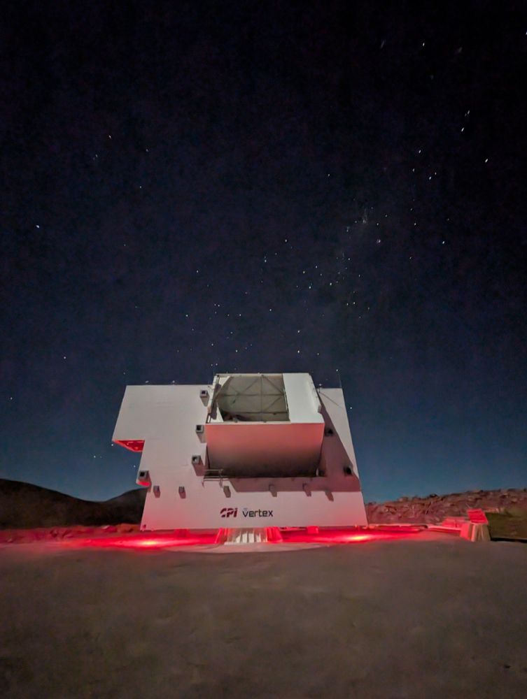 The Simons Observatory LAT with its aperture open, looking at the sky at night. Red safety lights illuminate the bottom of the telescope structure, while a few stars shine in the background.
