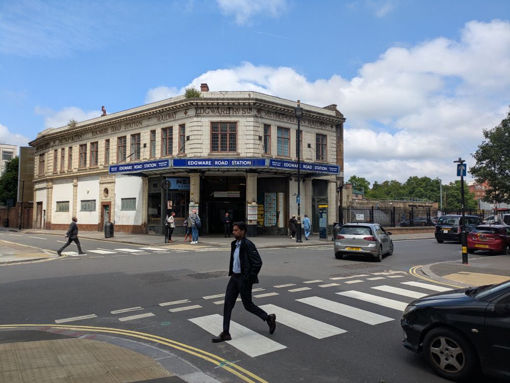 Junction of Chapel and Cabbell Streets. Two simple side road zebras without Belisha beacons can be seen with pedestrians crossing and a driver yielding. Edgware Road underground station entrance visible in the background.