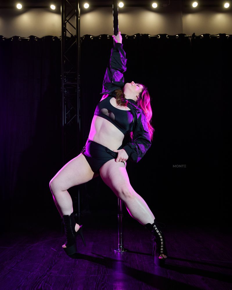 A woman posing in front of a pole, one hand grasping the pole and the other on her thigh as she looks up