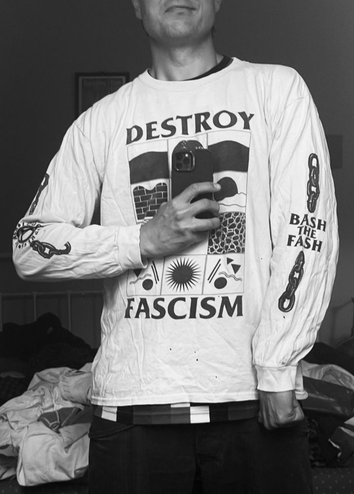 James wears a Black Lodge Press long sleeved T-Shirt with “DESTROY FASCISM” on the front, and “BASH THE FASH” on his left arm. He’s also got a Clapton CFC home shirt peeking out the bottom.
