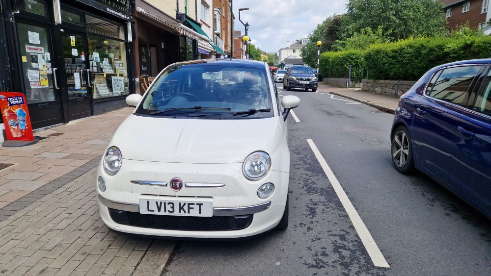 Small white car occupies part of the loading bay adjacent to small shops and also blocks a cycle lane, omcoming traffic fills the other lane. 