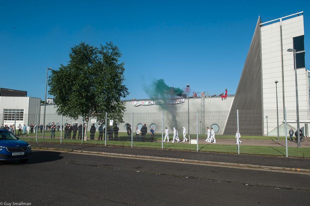 Image of Palestine Action protest at the Thale factory. They are on the roof setting off the kind of smoke flares commonly used in football stadiums. Thale staff can be seen casually walking past and standing nonchalantly around in the evacuation area in front of the building.