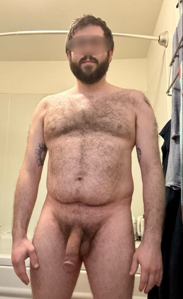 Hairy bearded tattooed bear cub man with messy hair from sleeping standing nude with a huge soft cock, arms to his side.