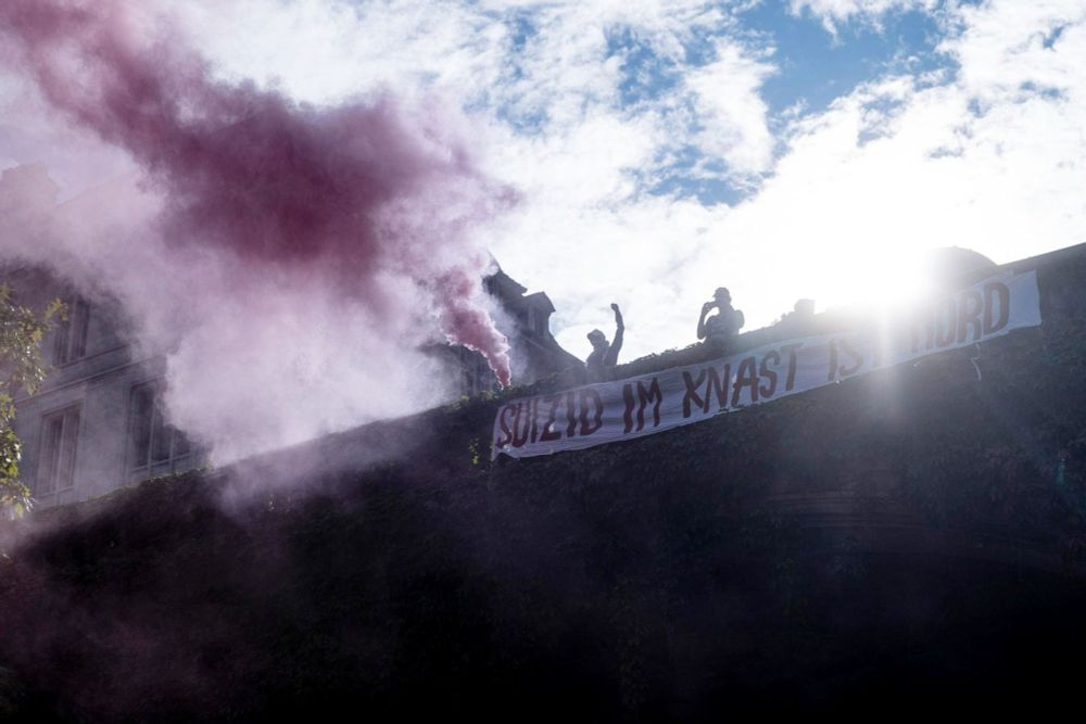 An einem Geländer auf einer Brücke ist ein Transparent angebracht: Suizid im Knast ist Mord. Auf der Brücke einige Personen mit einer Rauchpetarde.