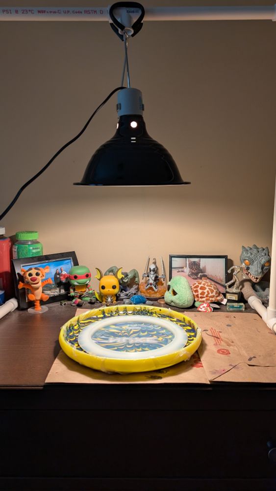 A picture of my heat lamp set up over a disc golf dye bed. The current disc that is being dyed is a Z Nuke. In the background are various tchotchkes, including Pops, pictures, and lots of turtles