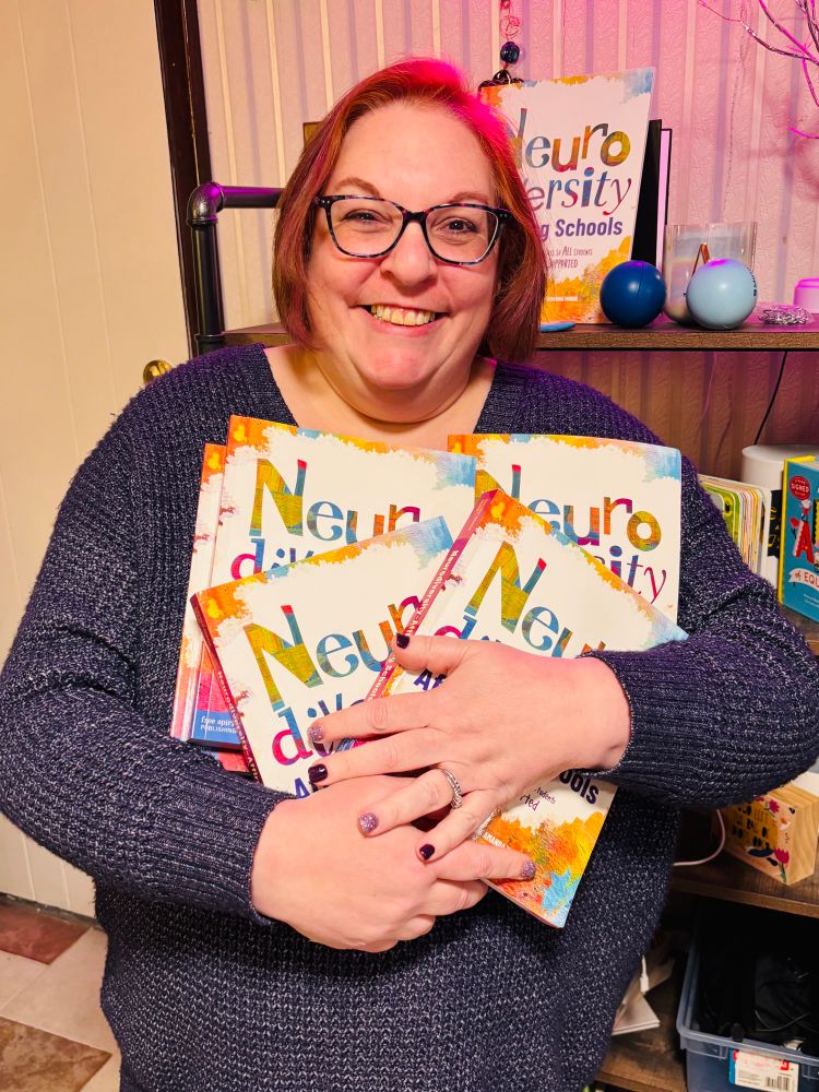 A smiling person with short red hair and glasses holds multiple copies of the book titled “Neurodiversity-Affirming Schools: Transforming Practices so All Students Feel Accepted & Supported.” They are wearing a dark purple sweater and standing in front of a bookshelf, with additional books and decorative items, including a colorful ball and illuminated tree, in the background.