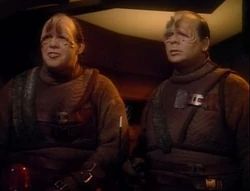 A photo of two pakled aliens. They look largely like fat white men, and are wearing clothes. They have facial prosthetics that create large vertical eyebrows, and slanting almond-shaped eyes. They have a confused affect on their face.