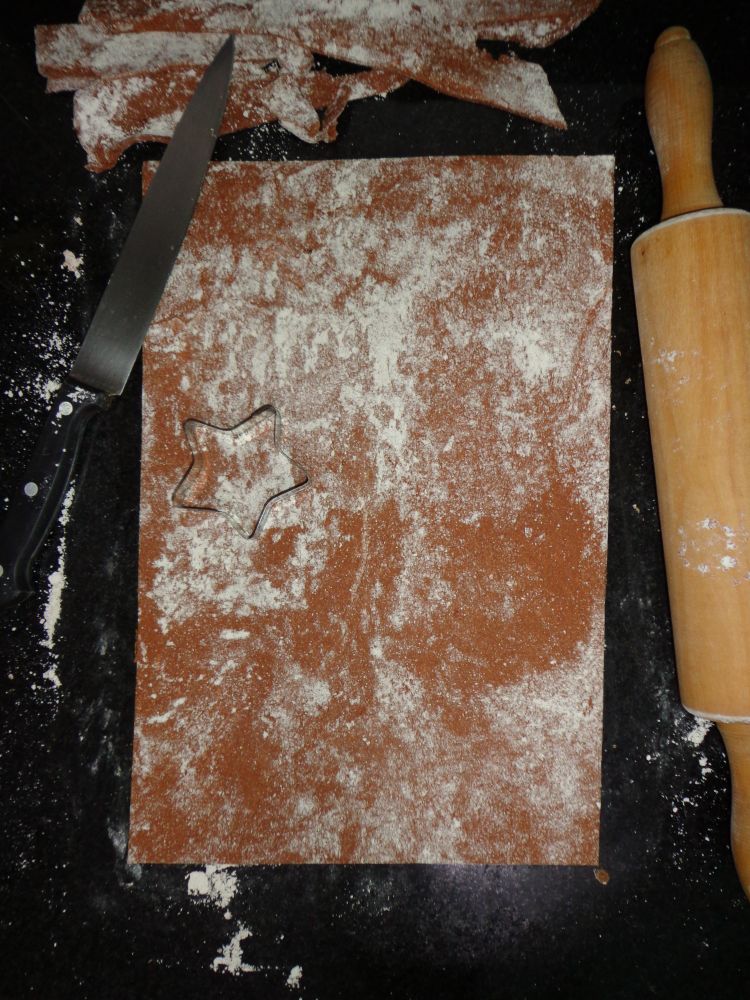 Apoiado sobre uma pedra preta, uma massa de petisco feita com fígado triturado, farinha de aveia, óleo de coco e pitada de canela aberta no rolo e cortada em um grande retângulo. Em cima da massa há um molde de metal de uma estrela, ao lado esquerdo há uma faca com rebarbas da mesma massa e, do lado direito, um rolo grande de madeira. Há farinha de aveia polvilhada na pedra e na massa.