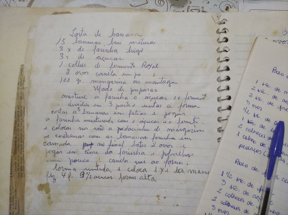 Caderno de folhas amareladas contendo a receita da torta de banana, no canto da imagem uma folha amarelada contendo a cópia de algumas receitas desse caderno e uma caneta bic azul. Transcrição da receita escrita: "Torta de Banana: 15 bananas bem maturas, 3x de farinha trigo, 3x de açúcar, 1 colher de fermento Royal, 2 ovos, canela em pó, 100 gr. margarina ou manteiga. Modo de preparo: misture a farinha, o açúcar e o fermento. Dividir em 3 partes e untar a forma. Cortar 5 bananas em fatoas e jogar a farinha mostira com açúcar e o fermento. E colocar no vão o pedacinho de margarina e continuar com as bananas, farinha em camada. No final bater 2 ovos e jogar em cima da farinha e polvilhar um pouco de canela, vai ao forno. [restante da transcrição, eu não compreendi]."