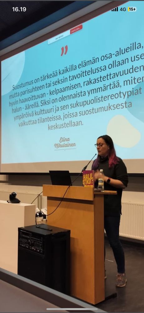 Kuva minusta ja kirjasta puhujanpöntön takana. Taustalla osa esitystäni,