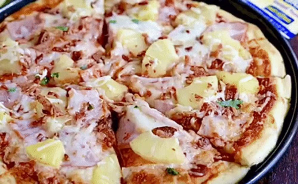 a pizza with pineapple , ham , cheese and bacon is sitting on a pan on a table .