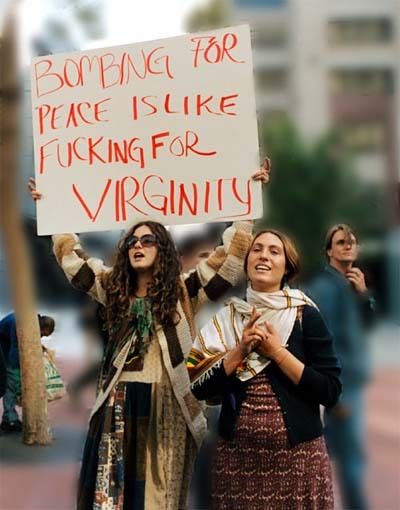 A photograph of two women demonstrators at an anti-war rally from the mid 2000s. One of them is holding up a sign that reads "Bombing for peace is like fucking for virginity"