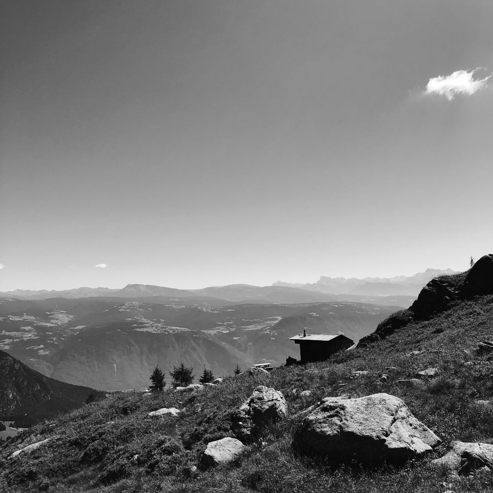 Un prato con dei sassi in montagna. Poco sotto una casetta e le cime delle Dolomiti in lontananza
