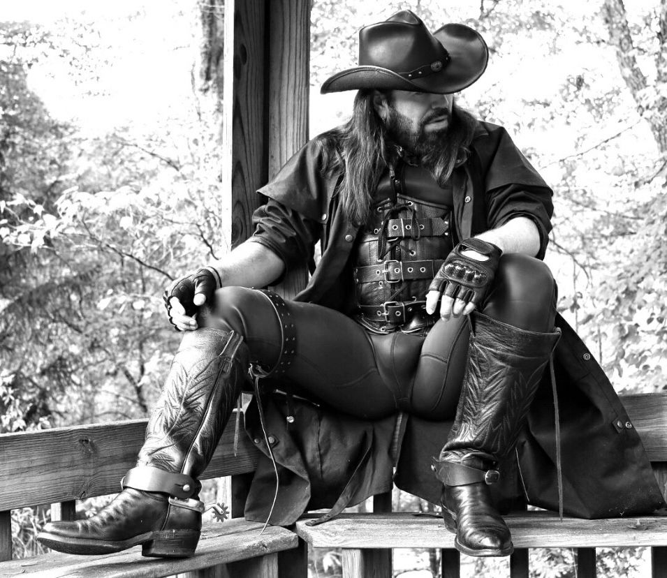 A male cowboy with long hair and a short beard, dressed in all black leather and tall boots, sits in a a wooden gazebo surrounded by trees.