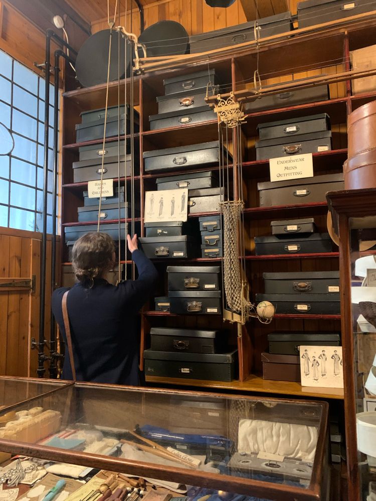 A woman seen from behind using a pulley system above shelves of boxes in a drapery shop 