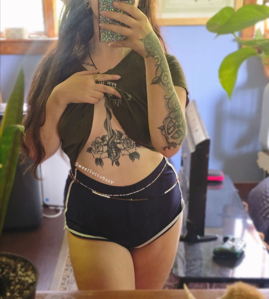 Viper is standing in front of a mirror and is wearing a green crop top tshirt and dark booty shorts. She is pulling the bottom of her shirt up to expose her sternum/underboob tattoo.
