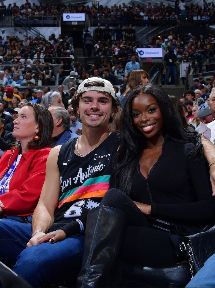 Olandria sitting courtside in a black outfit with black boots. Nic is sitting beside her in an San Antonio jersey. 