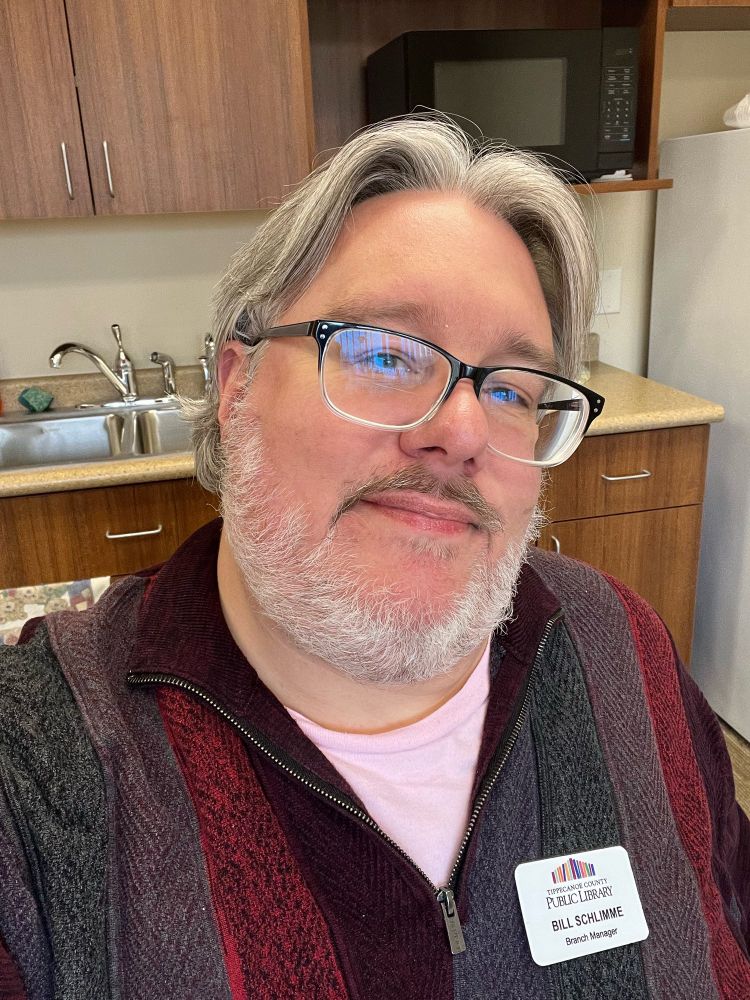 A middle aged man with glasses and a beard and hair long enough to curl at the back sitting in a break room at work.