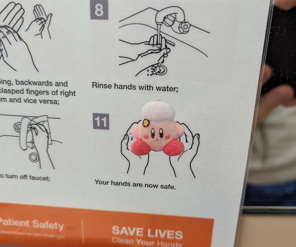 A handwashing instructional poster on a bathroom mirror. Only step 11 and surroundings are visible. Step 11 reads "Your hands are now safe" and has a drawn picture of hands holding a plush Kirby with a chef's hat on.