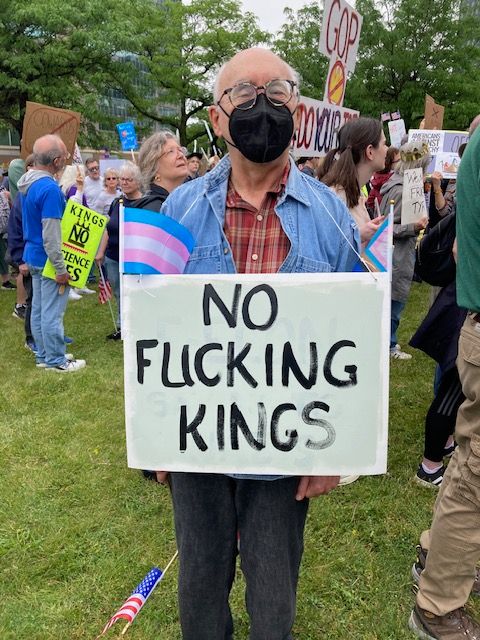 Cleveland, OH June 14th, 2025, No Kings Protest - Sorry about the language.
Description: Older man, wearing black KN95 mask, standing outdoors in crowd, holding sign that says NO F---ING KINGS, with Pride and Trans flags on sign, 