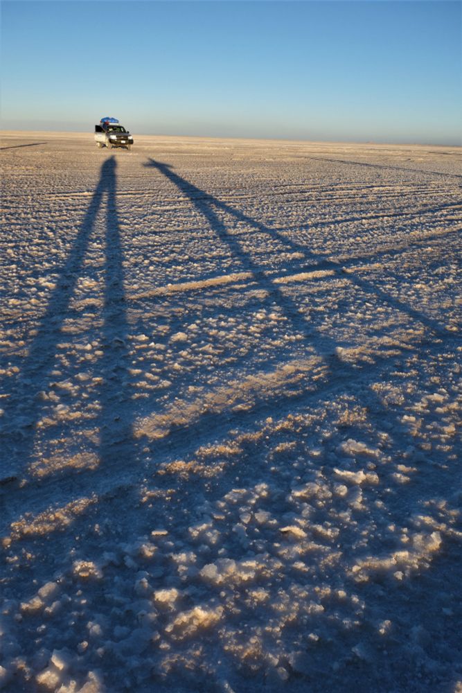 Extrem lange Schatten auf dem Boden der Salzwüste Uyuni, Bolivien. Tief stehende Sonne macht aus normalgewachsenen Menschen Kreaturen mit ultralangen Beinen. Blauer Himmel und ein Jeep am Horizont.