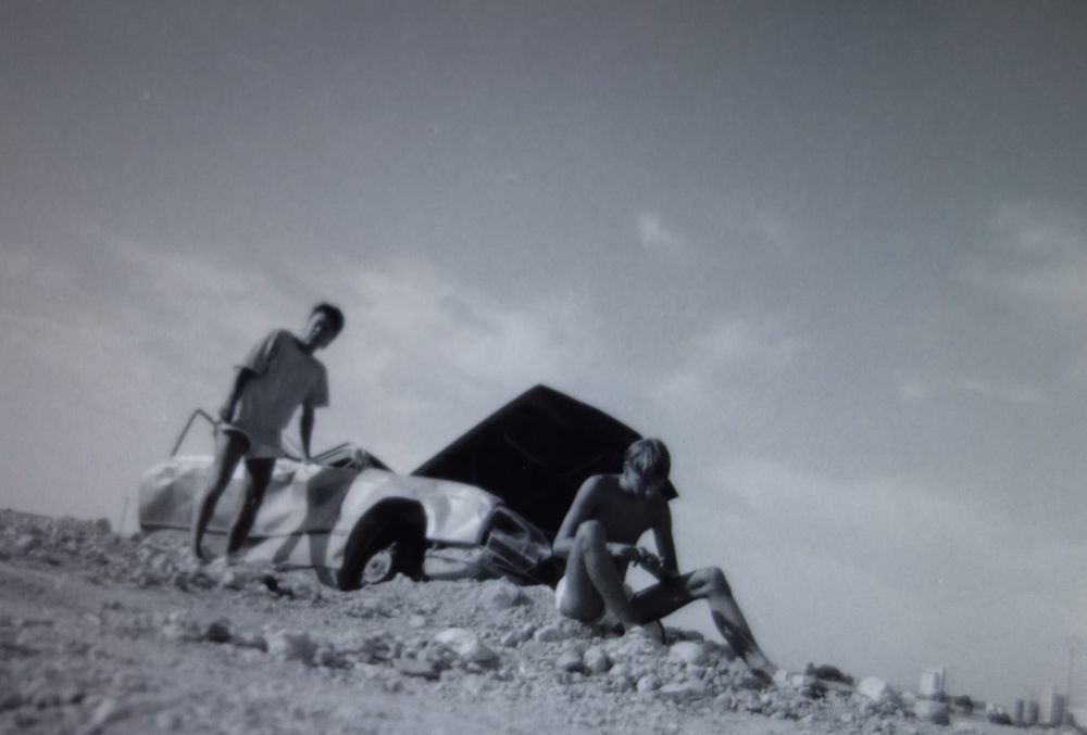 Schwarz Weiß Foto aus Bodenperspektive, Völlig verbeultes Autowrack, ohne Dach, Motorhaube halb hoch geklappt. Davor sitzt ein junger Mann auf sandig-schotterigem Boden und feilt sich die Fingernägel. Ein zweiter Mensch steht und stützt sich auf die Beifahrertür ab. Klarer Himmel mit wenigen Schleierwolken. 