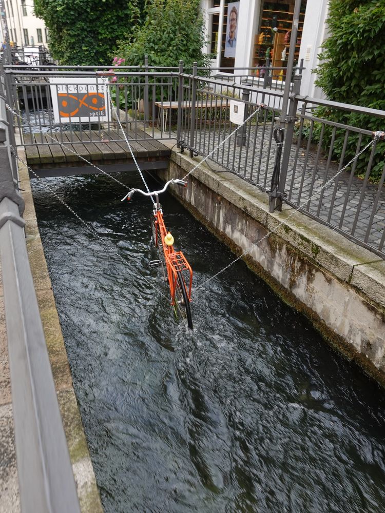 Orangefarbenes Fahrrad, das über fünf Ketten festgehalten knapp über einem Lech-Kanal in Augsburg hängt. Die Pedale werden durch das fließende Wasser angetrieben. Kunstaktion in der Nähe des Brecht-Geburtshauses.