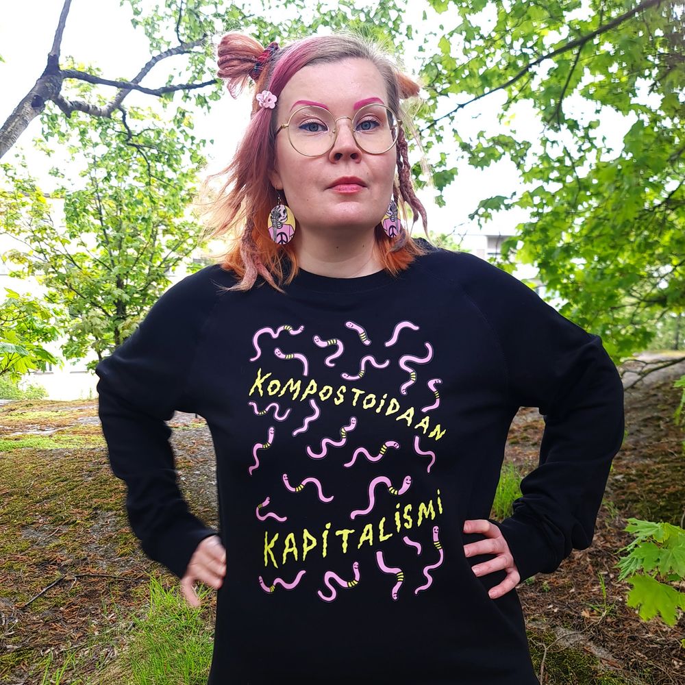 Satu kädet lanteilla päällään salaisuustason kompostoidaan kapitalismi pitkähihainen paita. Paidassa iloisia matoja, ja printti Kompostoidaan Kapitalismi