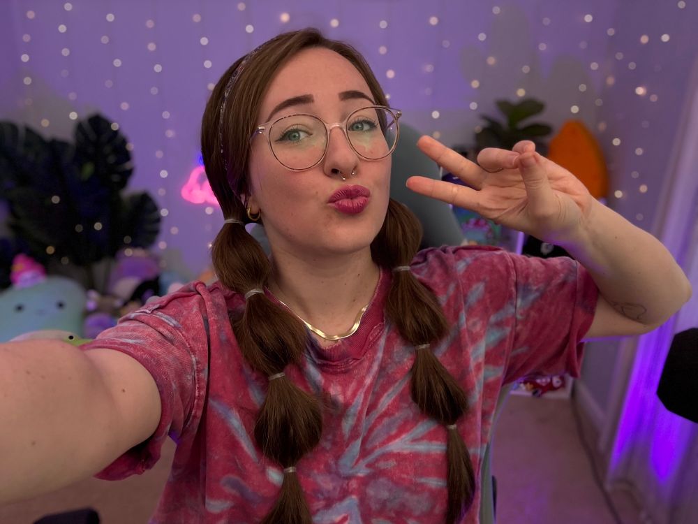 soph is wearing a pink tiedye shirt, bubble braids, and has pink lipstick on! she’s taking a selfie in front of her stream background 