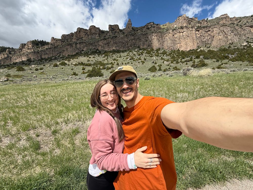 sophie and her partner are standing in a big grassy field with a giant cliff behind them, the photo is taken in a wide angle so the landscape and sky are pictured