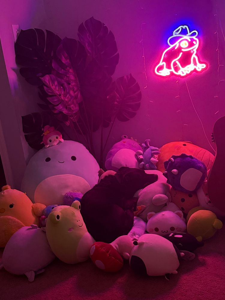 sophie’s dog tucker is laying on top of a huge pile of plushies with a bright pink frog neon light shining in the background 