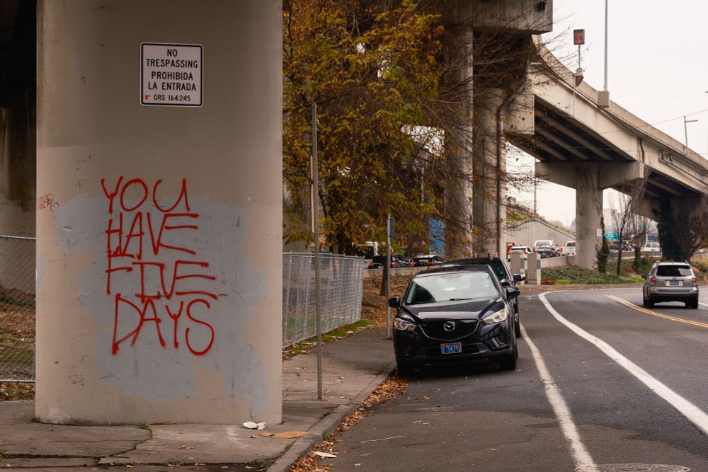 "YOU HAVE FIVE DAYS" spray painted on a pillar supporting the freeway in SE Portland. Above the graffiti, a sign from ODOT warning trespassers not to enter. Cars are parked on the street beside the pillar.
