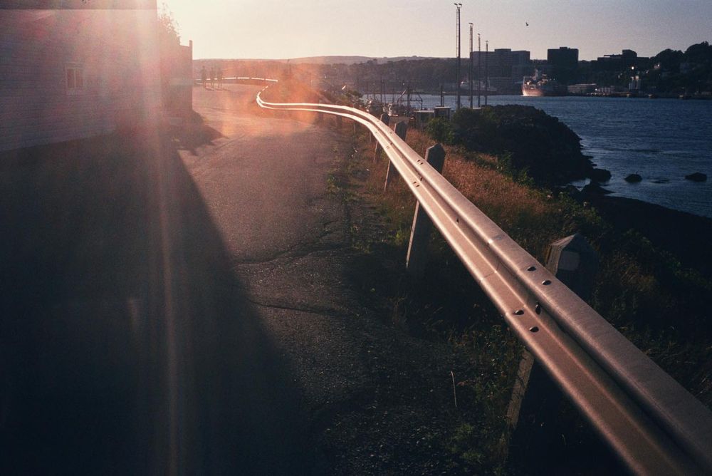 A 35mm film photo of a road winding along the edge of a harbour, with a light flare obscuring the end of the road and a city in the distance.