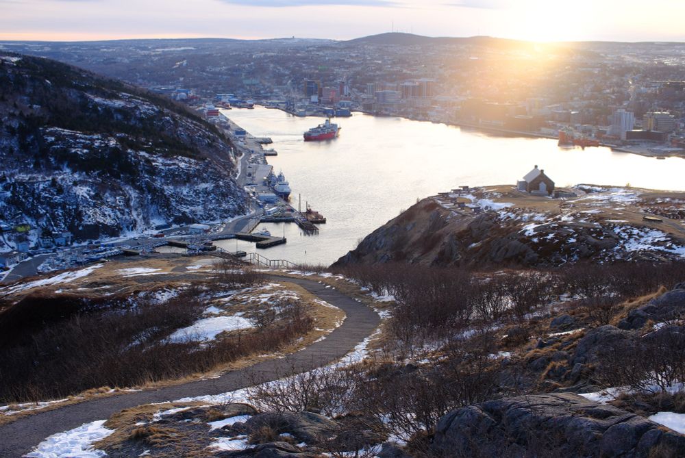 A view of St. John’s Harbour from Signal Hill, Newfoundland. A bit of snow along the edges of a walking path. The city sprawling along the sides of the harbour. A gold-tinged lens flare.