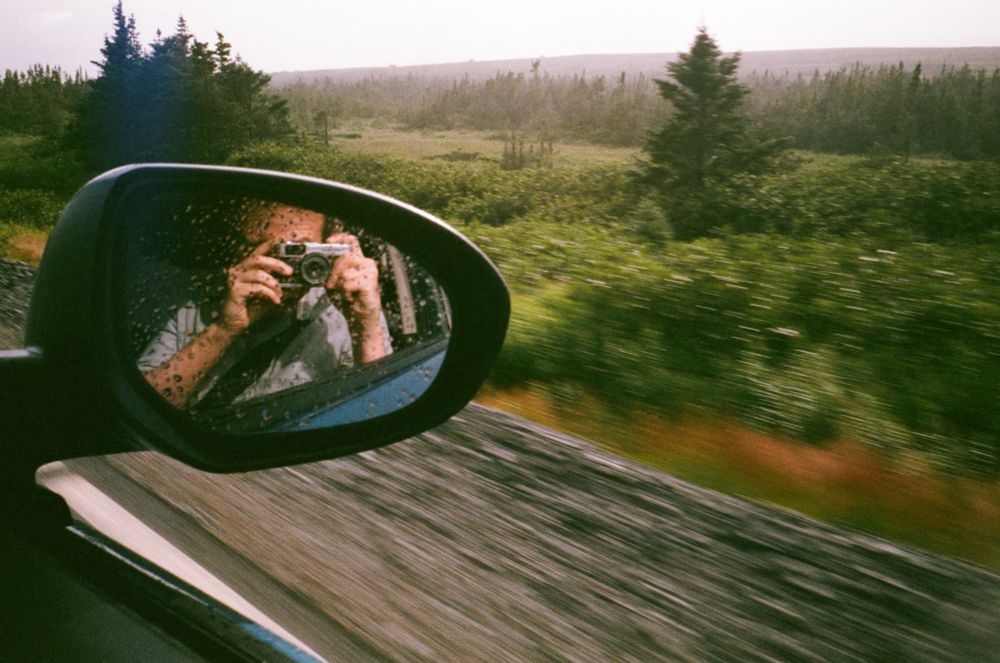 A retro-looking film photo taken from the passenger window of a moving car. The photographer is visible in the rear-view mirror, with a blurry landscape with trees and hills in the background.