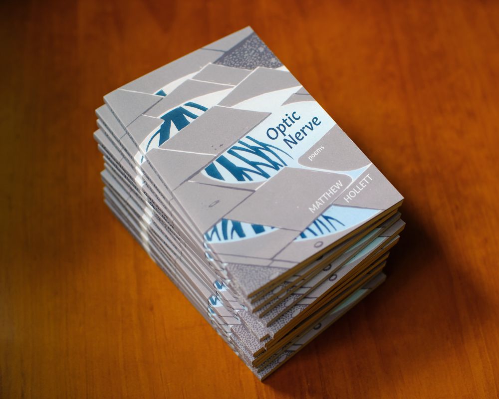 Several copies of a poetry collection titled "Optic Nerve", sitting on a wooden table. The book's cover image is an almost abstract graphical illustration of a tree reflected in a puddle shaped by paving stones.