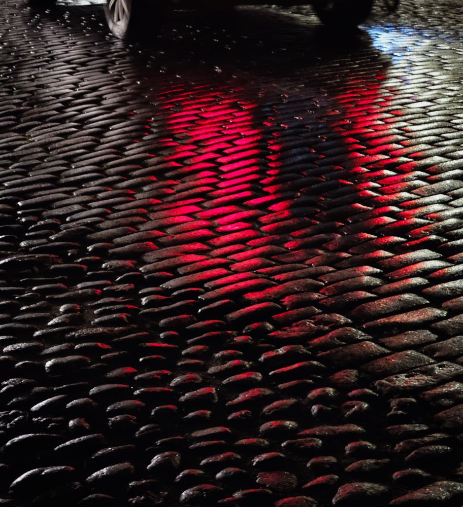 A cobblestone street at night, rounded stones shining bright red from the taillights of a passing car.