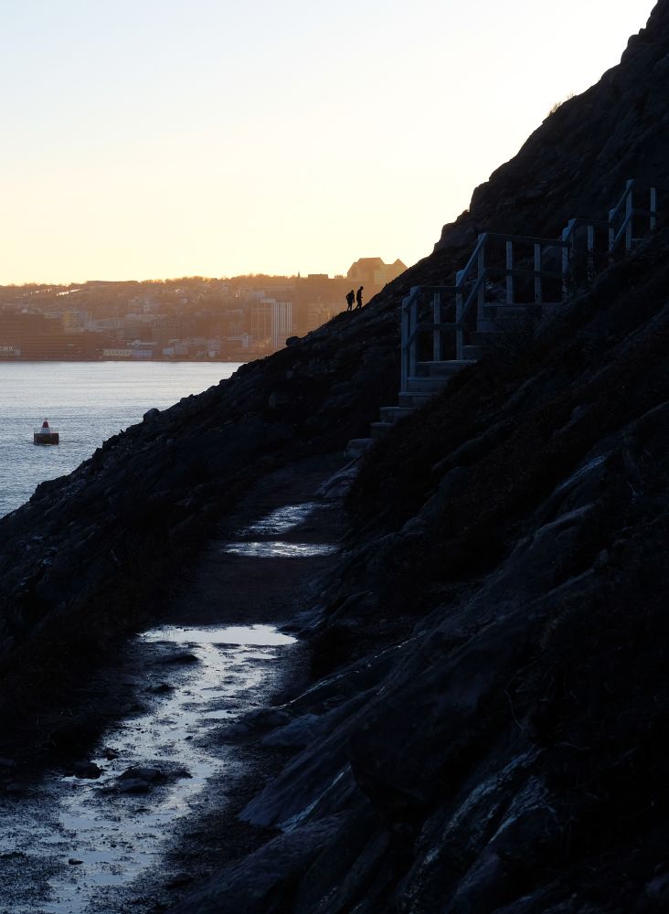 A hillside trail in deep blue shadow, with two hikers silhouetted in the distance. In the background is a port city glowing in warm evening light.