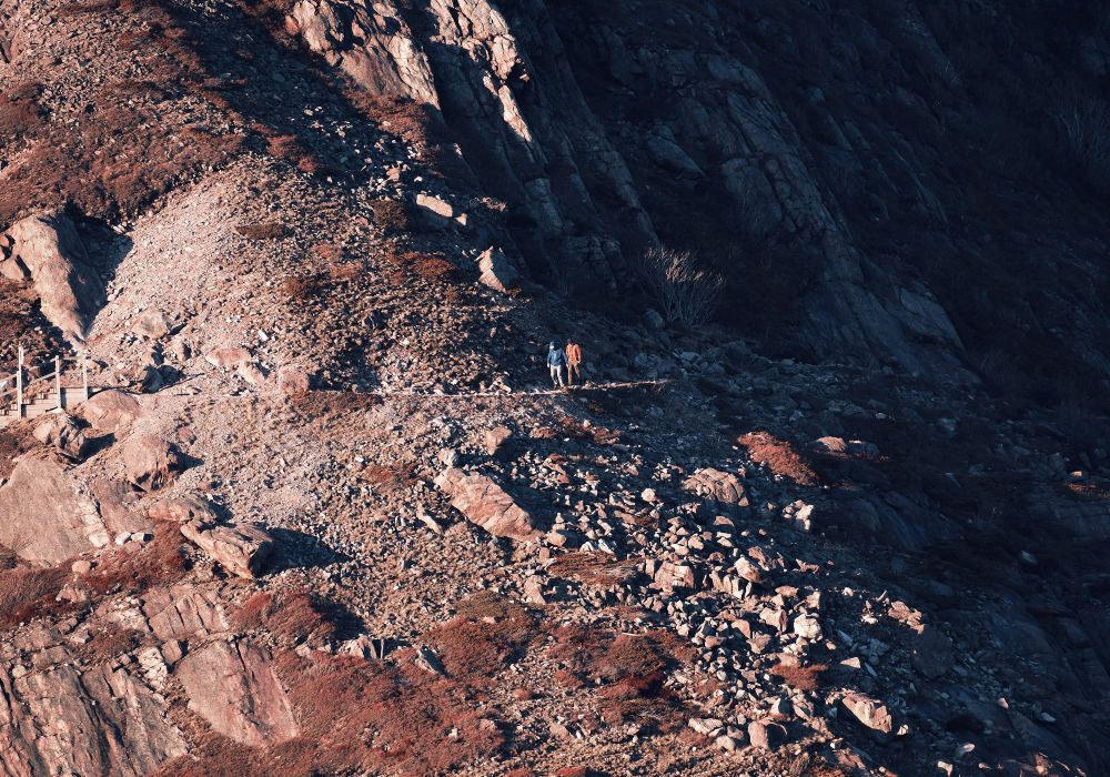 Two distant hikers on a hillside hiking trail, with dramatic evening light bringing out details in the rocky hill.