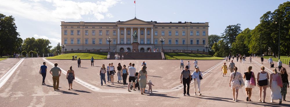 People walking on a plaza in front of a large, neoclassical building with a central statue. The area is surrounded by trees and open sky.