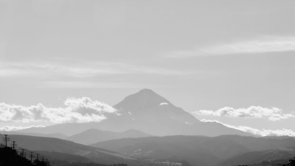 A black and white image of a mountain peak surrounded by clouds, with rolling hills and power lines in the foreground.