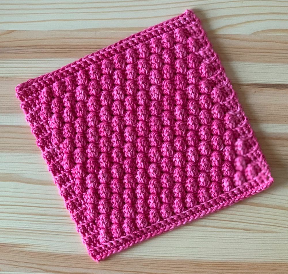 Textured pink crochet dishcloth laying on a pine table 