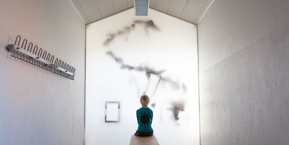 At the end of a school corridor, a boy sits on a bench looking at a giant dinosaur skeleton.

This is an artwork by american artist Michael McGillis.