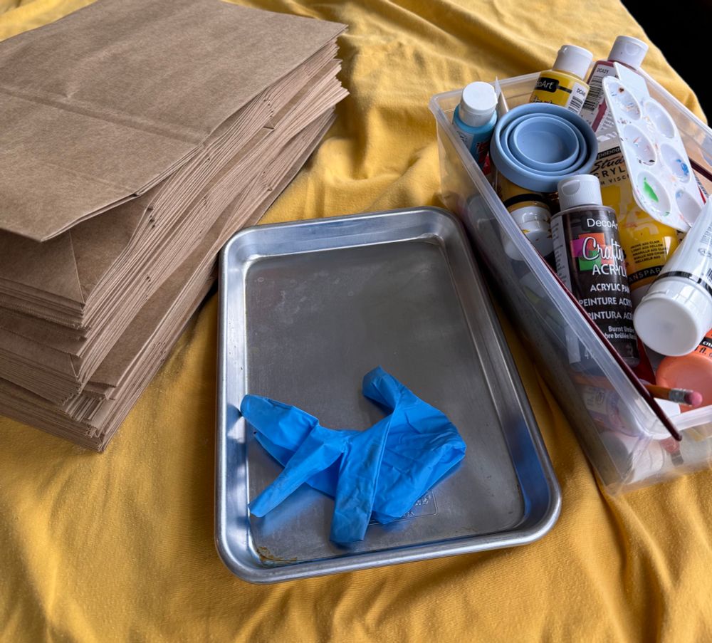 Blue glove in tray between stack of paper bags & acrylic paint supplies on yellow table cover 