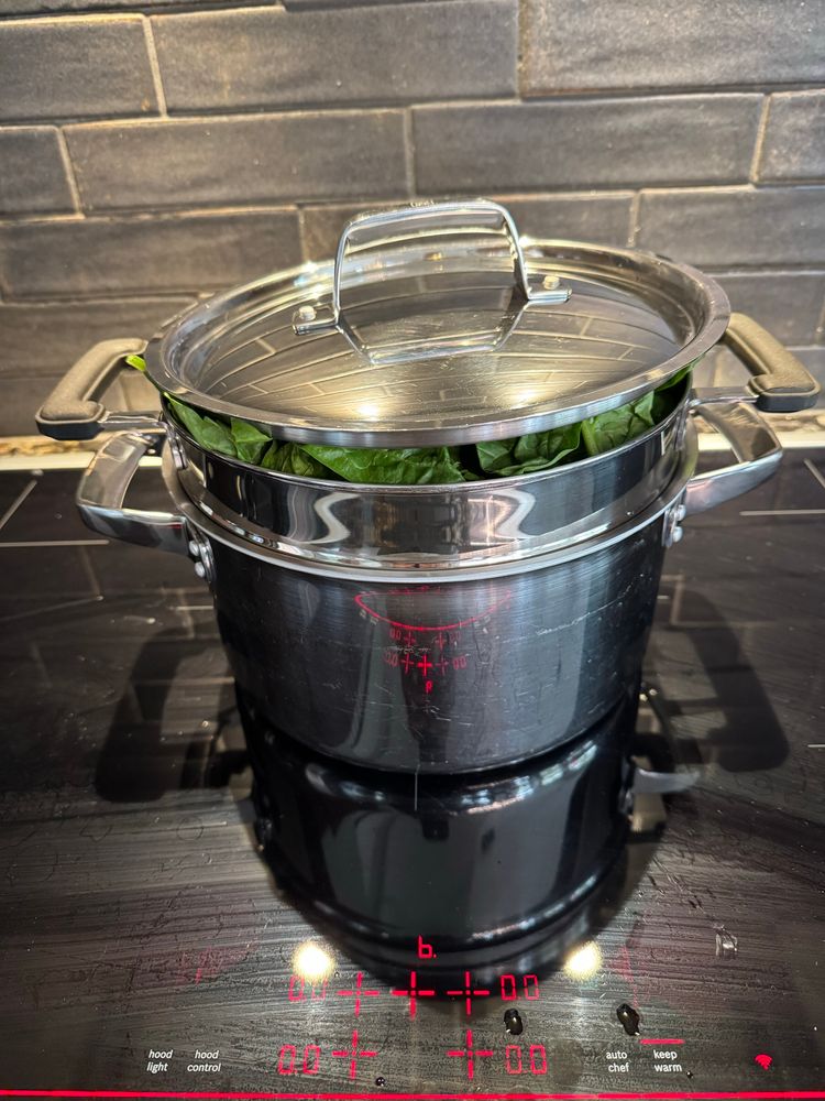 Lid skwishing down heaping pot of spinach on cooktop 