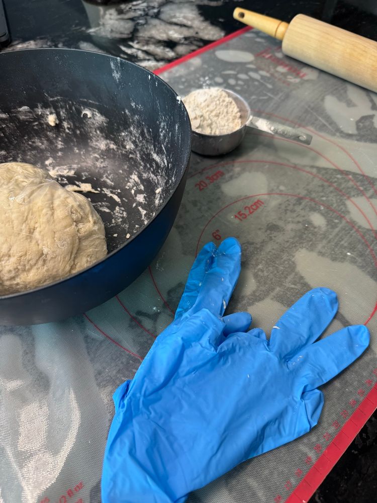 Dough wrapped in plastic in black bowl with blue nitrile gloves, rolling pin & flour on a white silicone mat.