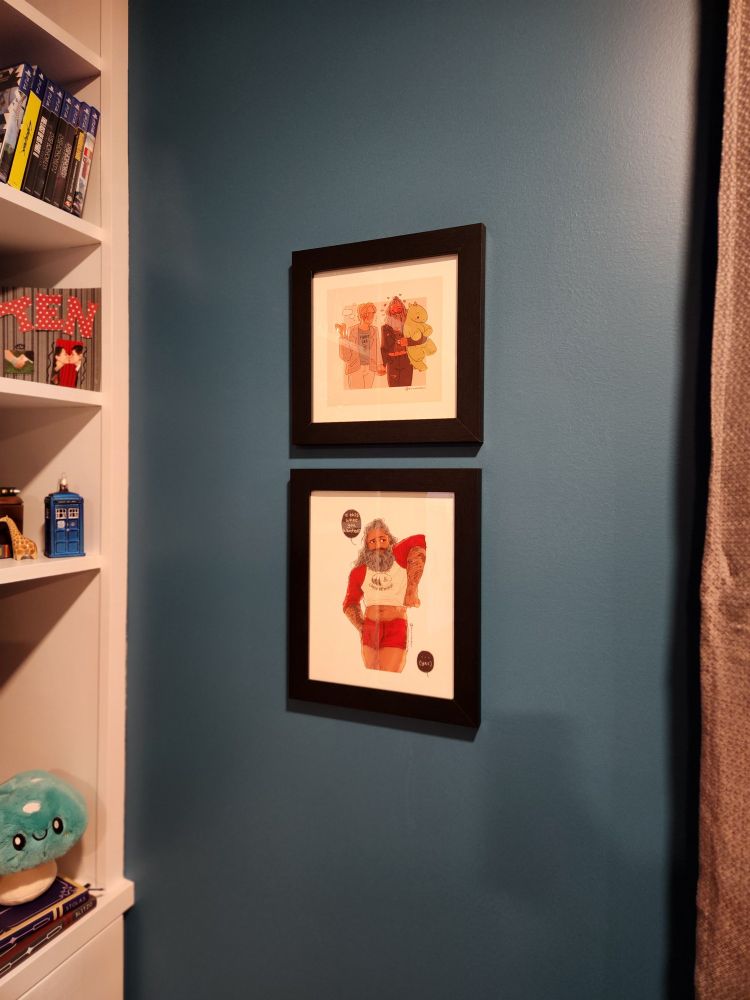 2 pieces of framed art based on two fanfics are hung on a teal wall. A shelf with toys and video games is on the left, grey curtain on the right.
In the top piece of art, Stede and Ed are walking side by side, Ed is holding an overstuffed dinosaur that was won in a fair game. Ed's wearing black jeans, a black leather jacket, and a Metallica shirt that is short enough to show his fuzzy tummy; Stede is wearing a plaid blazer, khakis, and a t-shirt that says "plant dad" the two are holding hands and smiling. 
In the bottom piece, Ed is shown wearing short red shorts and a raglan 3/4 sleeve shirt with red sleeves and white body that is too small, exposing his fuzzy tummy. The shirt says "camp revenge" with a small picture of trees, a lake, and a boat. Ed has a cute, coy look on his face and he's saying "is this what you wanted?" Ed has a noticable bulge in his shorts.